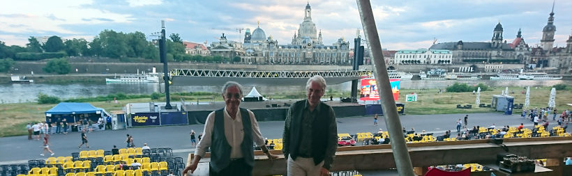 Guido und Maurizio stehen oben auf der Tribüne und schauen in die Kamera, im Hintergrund unten die Bühne, dahinter die Elbe und die Silhouette der Stadt Dresden